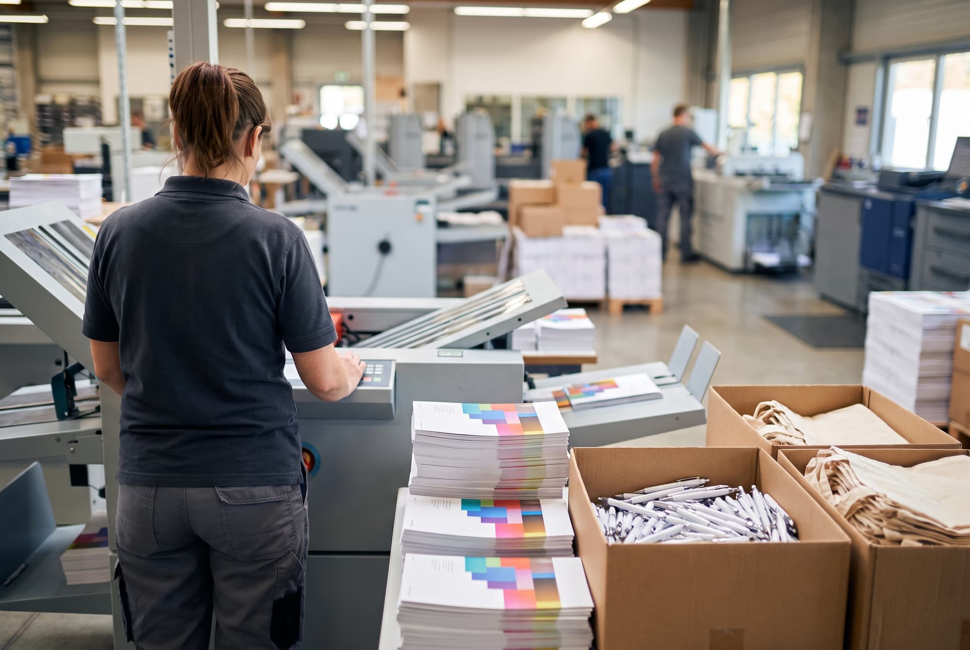 Stapel gedruckter Broschüren und Kartons mit Kugelschreibern und Stofftaschen in einer Druckerei